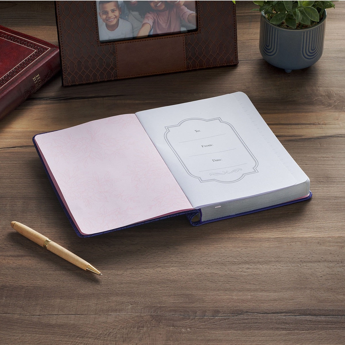Open purple faux leather journal with pen on wooden surface next to framed photo and potted plant