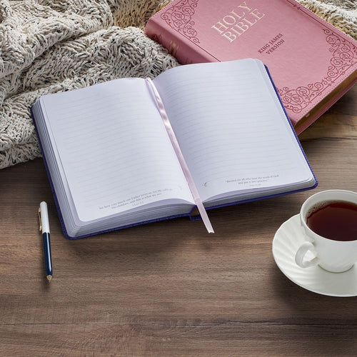 Purple faux leather journal open to lined pages with ribbon marker, pen beside, pink Bible and tea cup on wooden table